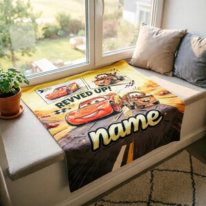 May include: A yellow and brown blanket featuring Lightning McQueen and other characters from the movie Cars. The blanket has the words "REVVED UP!" and "name" printed on it. The blanket is displayed on a window seat.