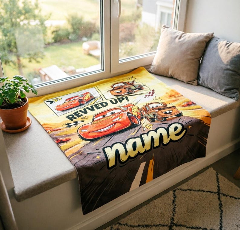 May include: A yellow and brown blanket featuring Lightning McQueen and other characters from the movie Cars. The blanket has the words "REVVED UP!" and "name" printed on it. The blanket is displayed on a window seat.