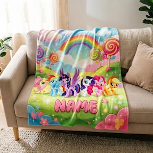 May include: A soft fleece blanket featuring a colourful My Little Pony graphic with six characters, a bright rainbow, and large lollipops against a green landscape. The word NAME is printed in large pink letters at the bottom of the blanket.