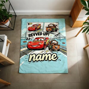 May include: A light blue blanket featuring cartoon images of the Cars movie characters. The blanket has a comic strip design with the text "REVVED UP!" and the word "name" in a stylized font. The main characters are Lightning McQueen and Mater.