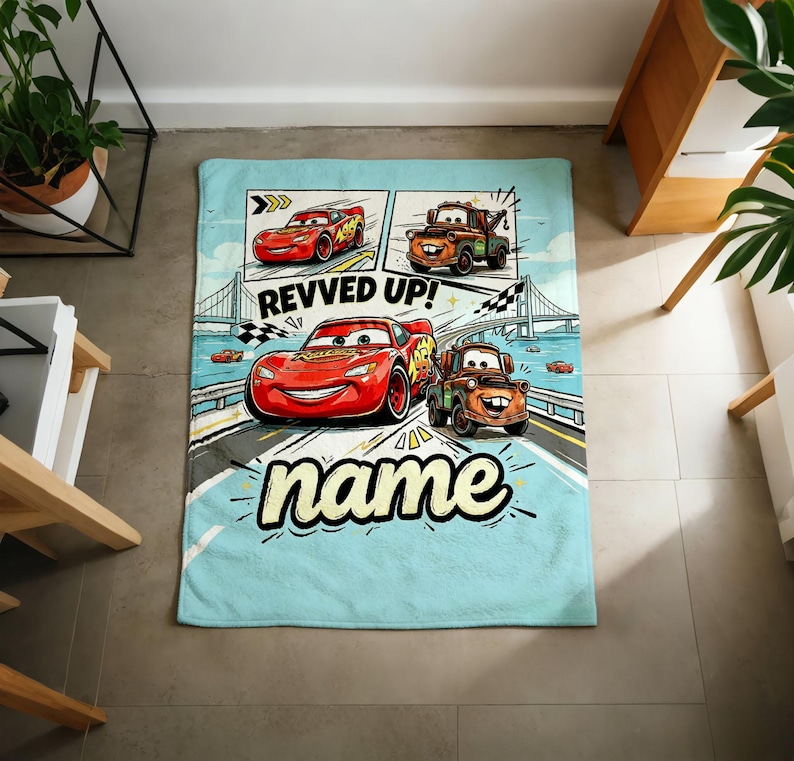 May include: A light blue blanket featuring cartoon images of the Cars movie characters. The blanket has a comic strip design with the text "REVVED UP!" and the word "name" in a stylized font. The main characters are Lightning McQueen and Mater.