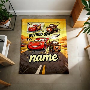 May include: A rectangular, yellow and brown blanket featuring cartoon race cars. The blanket has the text "REVVED UP!" and "name" in a bold font. The design includes images of red and brown race cars on a racetrack.