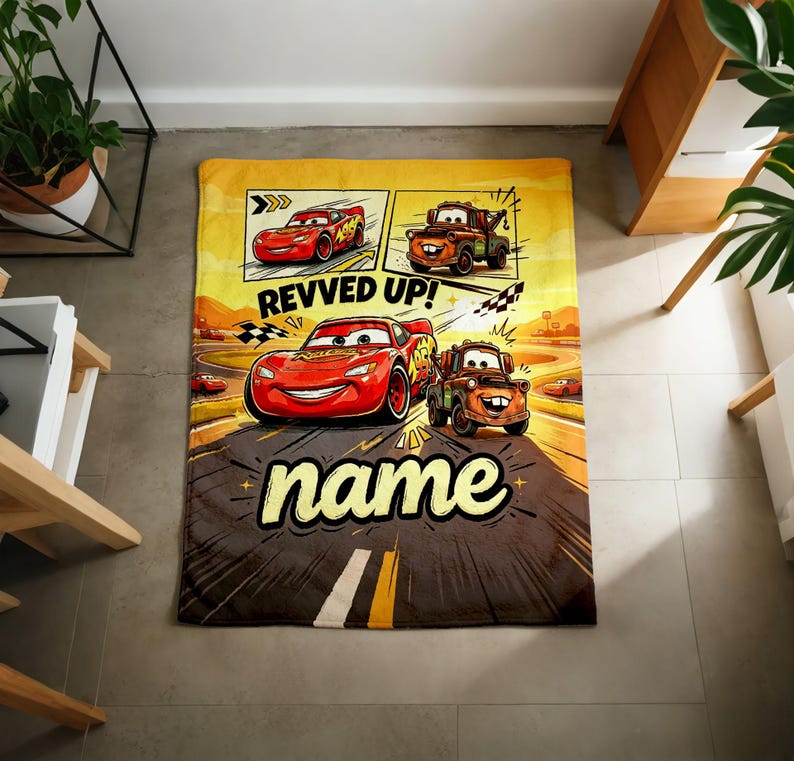 May include: A rectangular, yellow and brown blanket featuring cartoon race cars. The blanket has the text "REVVED UP!" and "name" in a bold font. The design includes images of red and brown race cars on a racetrack.