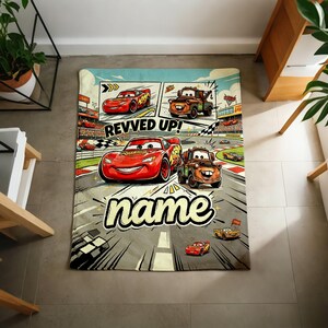 May include: A rectangular blanket featuring a comic-style design with the characters Lightning McQueen and Mater from the movie Cars. The blanket has a racing theme with the text "REVVED UP!" and "name" in a playful font. The color palette includes red, orange, and gray.