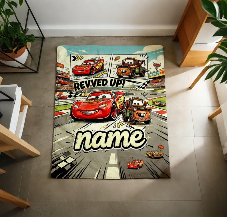 May include: A rectangular blanket featuring a comic-style design with the characters Lightning McQueen and Mater from the movie Cars. The blanket has a racing theme with the text "REVVED UP!" and "name" in a playful font. The color palette includes red, orange, and gray.