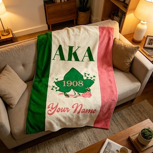 Op de afbeelding: Een zachte, crèmekleurige deken met groene en roze strepen. De deken heeft de tekst "AKA" in groen, een groen klimopbladontwerp met "1908" in het midden en de woorden "Your Name" in roze. De deken is over een beige bank gedrapeerd.