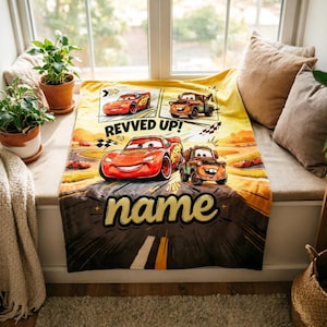 May include: A yellow and brown blanket featuring Lightning McQueen and Mater from the Cars movie. The blanket has the words "REVVED UP!" and "name" printed on it. The blanket is displayed on a window seat.