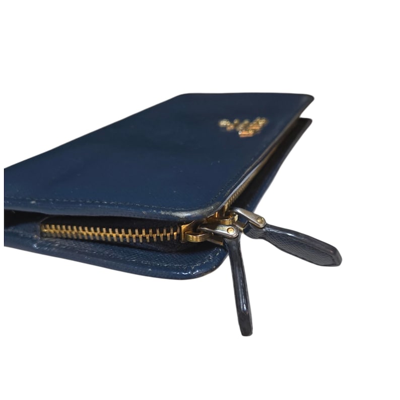 May include: A navy blue leather wallet with a gold-colored zipper and a small gold logo. The wallet is rectangular and has a glossy finish. The zipper pull is also navy blue.