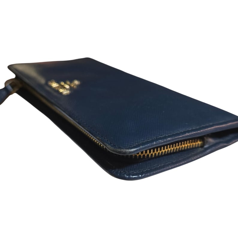 May include: A navy blue leather wallet with a gold zipper and a gold logo. The wallet is rectangular and has a textured surface. The zipper is open, revealing the interior.