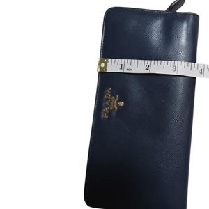 May include: A navy blue Prada wallet with a gold logo is measured with a tape measure. The wallet is approximately 4 inches wide. The wallet has a textured surface.