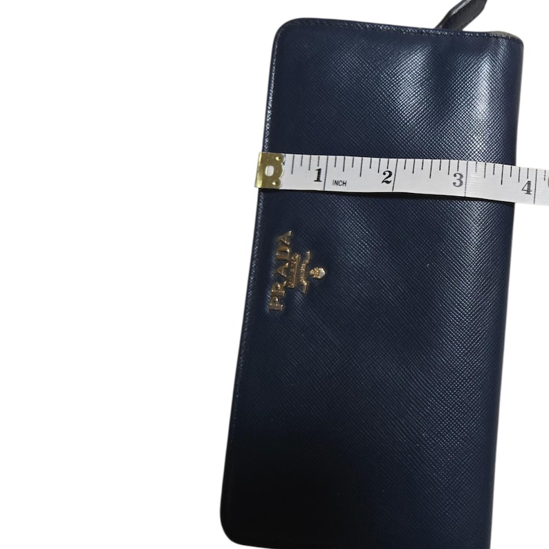 May include: A navy blue Prada wallet with a gold logo is measured with a tape measure. The wallet is approximately 4 inches wide. The wallet has a textured surface.