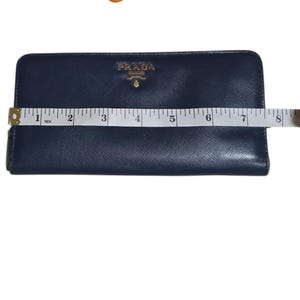 May include: A navy blue Prada wallet with a gold logo, measuring approximately 8 inches in length. The wallet is rectangular and has a textured surface. A measuring tape is placed across the wallet.