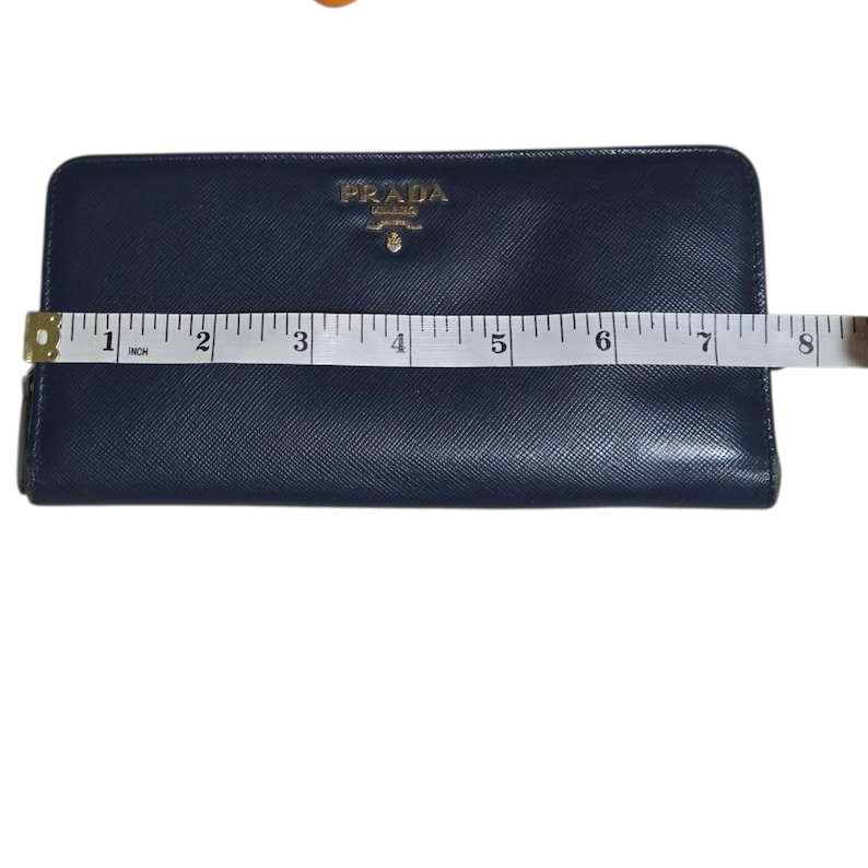 May include: A navy blue Prada wallet with a gold logo, measuring approximately 8 inches in length. The wallet is rectangular and has a textured surface. A measuring tape is placed across the wallet.