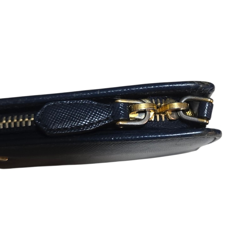 May include: Close-up of a navy blue wallet with a gold-colored zipper. The wallet has a textured surface and a gold zipper pull. The zipper is partially open, revealing the interior.
