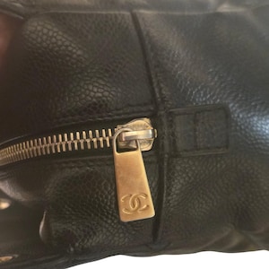 May include: Close-up of a black leather handbag with a gold-colored zipper. The zipper pull features the Chanel logo. The leather has a textured, pebbled appearance. The bag appears to be well-worn.