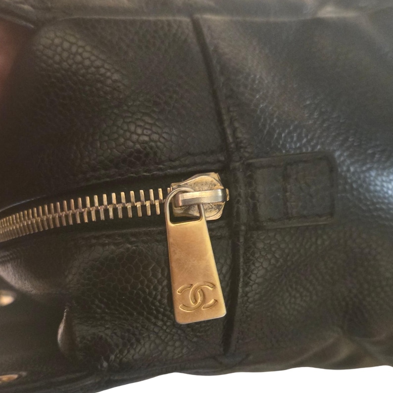 May include: Close-up of a black leather handbag with a gold-colored zipper. The zipper pull features the Chanel logo. The leather has a textured, pebbled appearance. The bag appears to be well-worn.