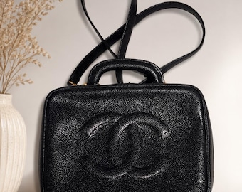 May include: A black rectangular handbag with a textured caviar leather finish and a prominent embossed interlocking CC logo on the front. The bag features a top handle and a long shoulder strap, with gold-tone hardware visible at the strap attachments.