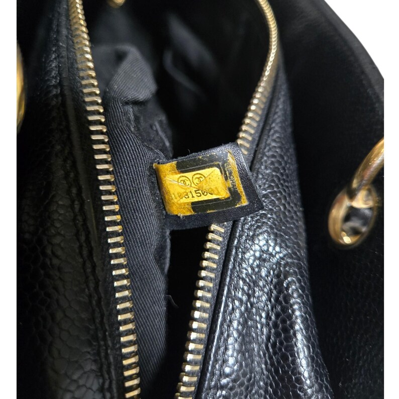 May include: Close-up of a black leather handbag with a gold zipper and hardware. A yellow tag with the number 18315601 is visible. The bag has a textured surface and a black interior.