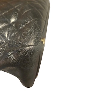 May include: Close-up of a black, quilted leather handbag. The bag features a diamond pattern and a textured surface. A gold-colored metal accent is visible on the corner of the bag. The bag is set against a white background.