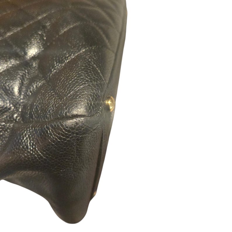 May include: Close-up of a black, quilted leather handbag. The bag features a diamond pattern and a textured surface. A gold-colored metal accent is visible on the corner of the bag. The bag is set against a white background.