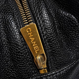May include: Close-up of a black leather bag with a gold-colored zipper. The zipper pull features the brand name "CHANEL" in gold lettering. The leather has a textured, pebbled appearance.