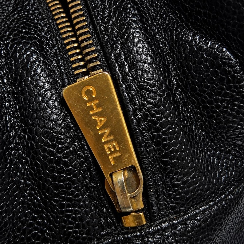 May include: Close-up of a black leather bag with a gold-colored zipper. The zipper pull features the brand name "CHANEL" in gold lettering. The leather has a textured, pebbled appearance.