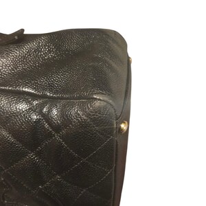 May include: Close-up of a black leather handbag with a quilted diamond pattern. The bag has a textured surface and gold-colored metal feet. The corner of the bag is visible, showing wear.
