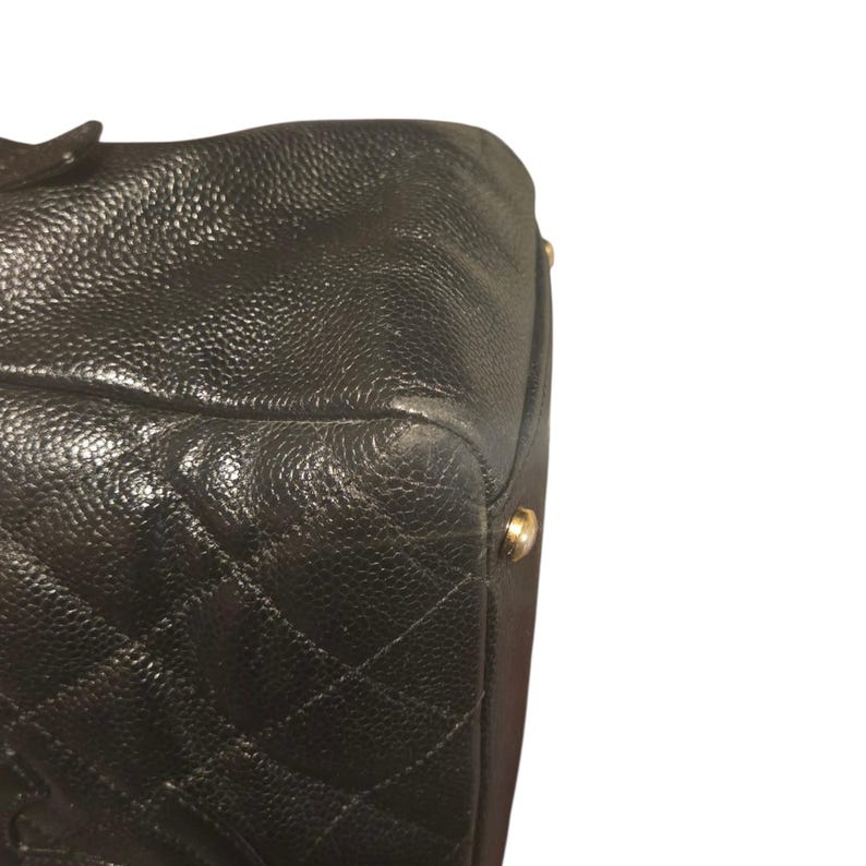 May include: Close-up of a black leather handbag with a quilted diamond pattern. The bag has a textured surface and gold-colored metal feet. The corner of the bag is visible, showing wear.