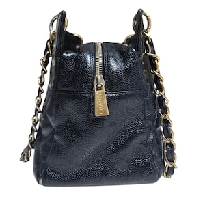 May include: Black leather handbag with gold-tone chain straps and hardware. The bag features a top zipper closure with a gold pull tab. The bag has a quilted design and a pebbled texture. The brand name "CHANEL" is visible on the zipper pull.