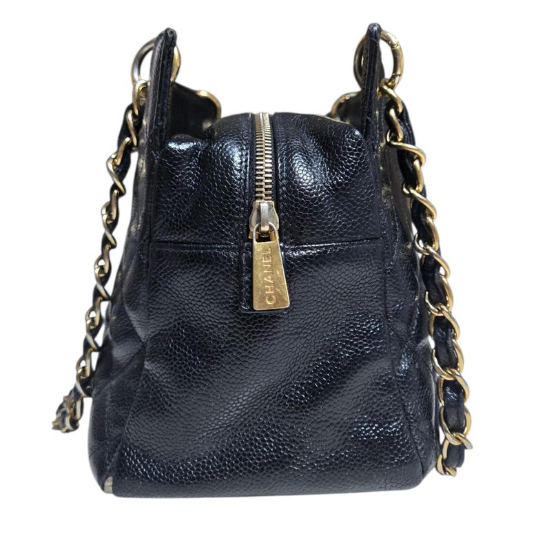May include: Black leather handbag with gold-tone chain straps and hardware. The bag features a top zipper closure with a gold pull tab. The bag has a quilted design and a pebbled texture. The brand name "CHANEL" is visible on the zipper pull.