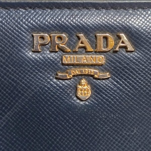 May include: Close-up of a navy blue leather Prada wallet. The gold-colored Prada logo is prominently displayed at the top, with "MILANO" and "DAL 1913" below. The wallet has a textured surface.