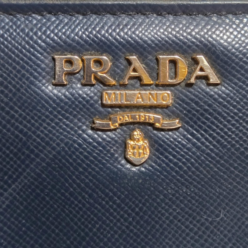 May include: Close-up of a navy blue leather Prada wallet. The gold-colored Prada logo is prominently displayed at the top, with "MILANO" and "DAL 1913" below. The wallet has a textured surface.