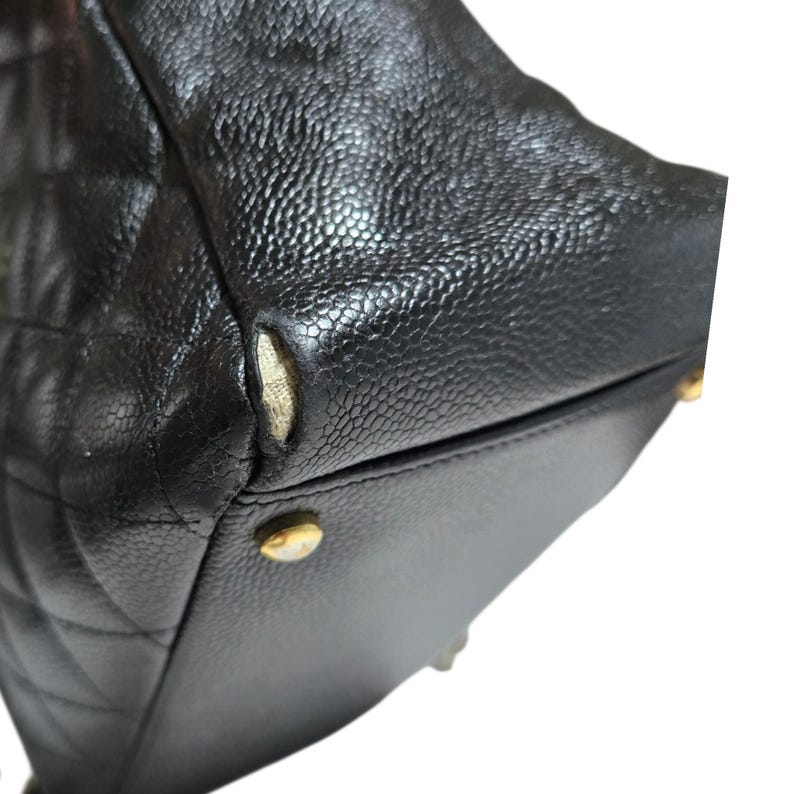 May include: Close-up of a black leather handbag with a diamond-quilted pattern. The bag shows wear and tear, with a visible tear on the corner. Gold-toned metal feet are visible on the bottom.