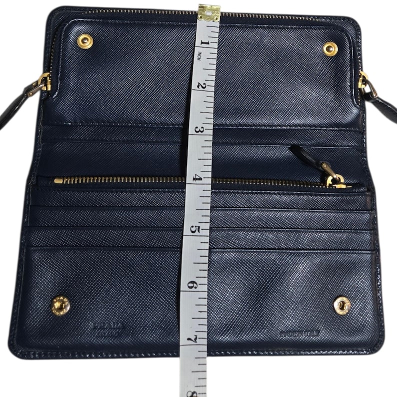 May include: Open navy blue wallet with gold-tone hardware, measuring approximately 7 inches in height. Features multiple card slots, a zippered compartment, and a snap closure. The brand name is visible. Made in Italy.