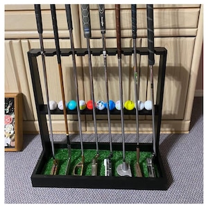 May include: A black wooden golf club rack holding several golf clubs and golf balls. The rack has a green turf base and a shelf for golf balls. The golf clubs are of various styles and colors, with black grips and silver shafts.