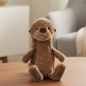 Op de afbeelding: Een bruin pluche otter speelgoed met een zacht, getextureerd uiterlijk. Het knuffeldier heeft een afgeronde kop, kleine oren en een donkerbruine neus. Het zit op een houten oppervlak.