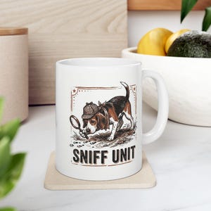 May include: White ceramic mug featuring a beagle detective with a magnifying glass and a plaid hat. The mug has the words "SNIFF UNIT" printed in a bold font. The design is framed with a decorative border.
