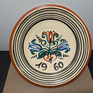 May include: A round, ceramic plate with a cream-colored center and a brown rim. The plate features a floral design in blue, green, and orange, with the number "1960" below the design. Black stripes encircle the center.