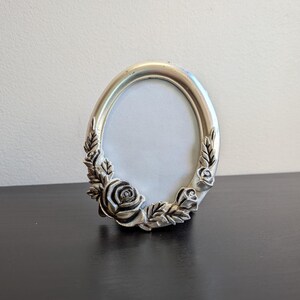 May include: An oval-shaped picture frame with a metallic silver finish. The frame features a raised, three-dimensional relief design of rose blossoms and leaves along the lower left and right sides, surrounding a plain white oval center insert.