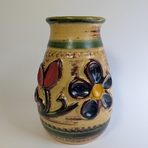 May include: A tan ceramic vase with a textured surface. The vase features a green rim and a green band near the top and bottom. It is decorated with a red tulip and a blue and yellow flower.