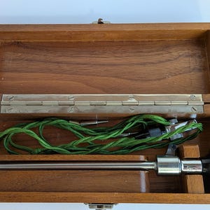 May include: Open wooden box with a hinged lid, containing a medical instrument. The instrument has a silver metal shaft, a black eyepiece, and a green cord. The box is made of a dark wood with a natural grain.