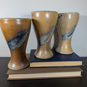 May include: Three tan ceramic goblets with a flared rim and a tapered base. Each goblet features a hand-painted blue whale illustration on the exterior. The glaze has a speckled, textured finish, and the vessels are arranged in a cluster.