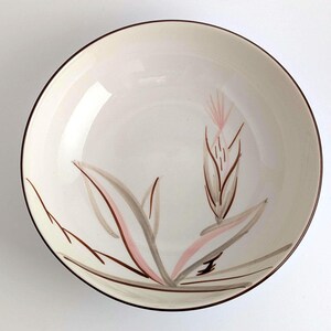 May include: A cream-colored ceramic bowl with a brown rim and a floral design. The design features stylized leaves and a central flower in shades of pink, brown, and gray. The bowl is round and appears to be vintage.