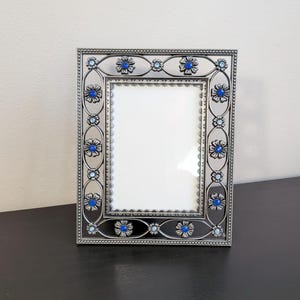 May include: A rectangular picture frame with a metallic silver finish, featuring an intricate openwork border decorated with small metal flowers and embedded blue and light blue rhinestones surrounding a central white rectangular display area.