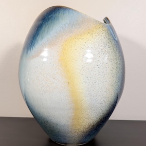 May include: A speckled ceramic vase with a unique, asymmetrical shape. The vase features a gradient of colors, including blue, yellow, and white. The top has a curved opening. The vase is a decorative item.