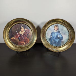 May include: Two circular brass plates featuring decorative embossed rims. The left plate displays a portrait of a person in a dark red outfit, while the right plate shows a portrait of a person in a blue outfit with a white ruffled collar.