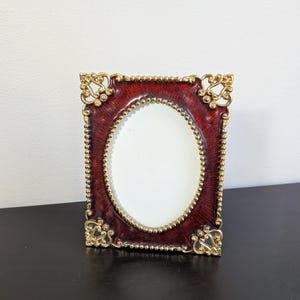 May include: A rectangular picture frame with a deep red, textured finish and an oval center opening. The frame features ornate gold-toned metal accents at each corner and a beaded gold-toned border surrounding the central oval display area.