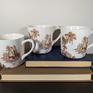 May include: Three white ceramic mugs with a brown and red pastoral scene design. The mugs are arranged on top of two books with blue and brown covers. The mugs have curved handles and are of varying sizes.