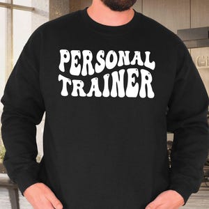 May include: Black crew neck sweatshirt with the words "PERSONAL TRAINER" in large, white, retro-style lettering. The sweatshirt is worn by a person in blue jeans, with hands in pockets. The background suggests a gym setting.