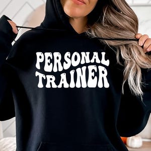 Op de afbeelding: Zwarte hoodie met de witte tekst "PERSONAL TRAINER" in een retro lettertype. De sweatshirt heeft een voorzak en een capuchon met trekkoord. De afbeelding toont een persoon die de hoodie draagt.